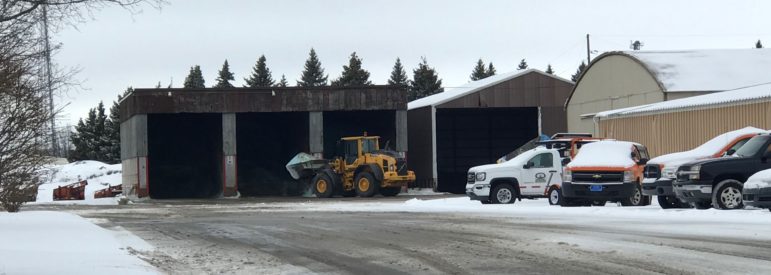 A snowy day, where there is a yellow construction vehicle scooping the salt up to place in a salt truck.