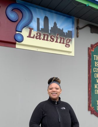 An African American woman in a black fleece smiles below a sign that reads "? Lansing"