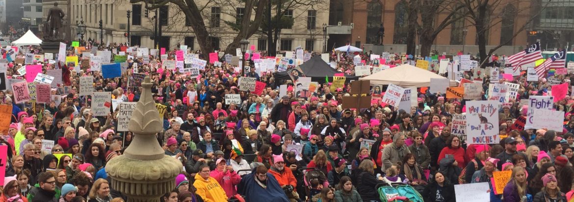 Hundreds of people, mostly women and many wearing pink hats, fill a plaza in front of the Michigan Capitol.
