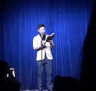 He is wearing a tan blazer and dark washed pants. He is reading his poem out of a journal and into a microphone. There is a blue curtain behind him.