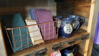 A shelf holds small goods that are sold at Blue Owl Coffee, including coffee mugs and journals.