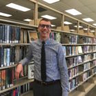 Scott Duimstra who is dressed in a blue button down shirt with a black tie leans on the shelves of books.