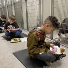 5 kids sit next to dog kennels and read books out loud.