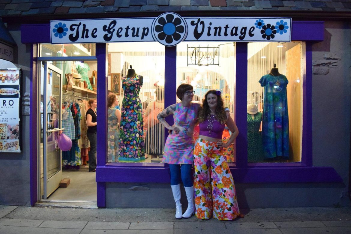 Two business partners in vintage clothes stand outside their store.