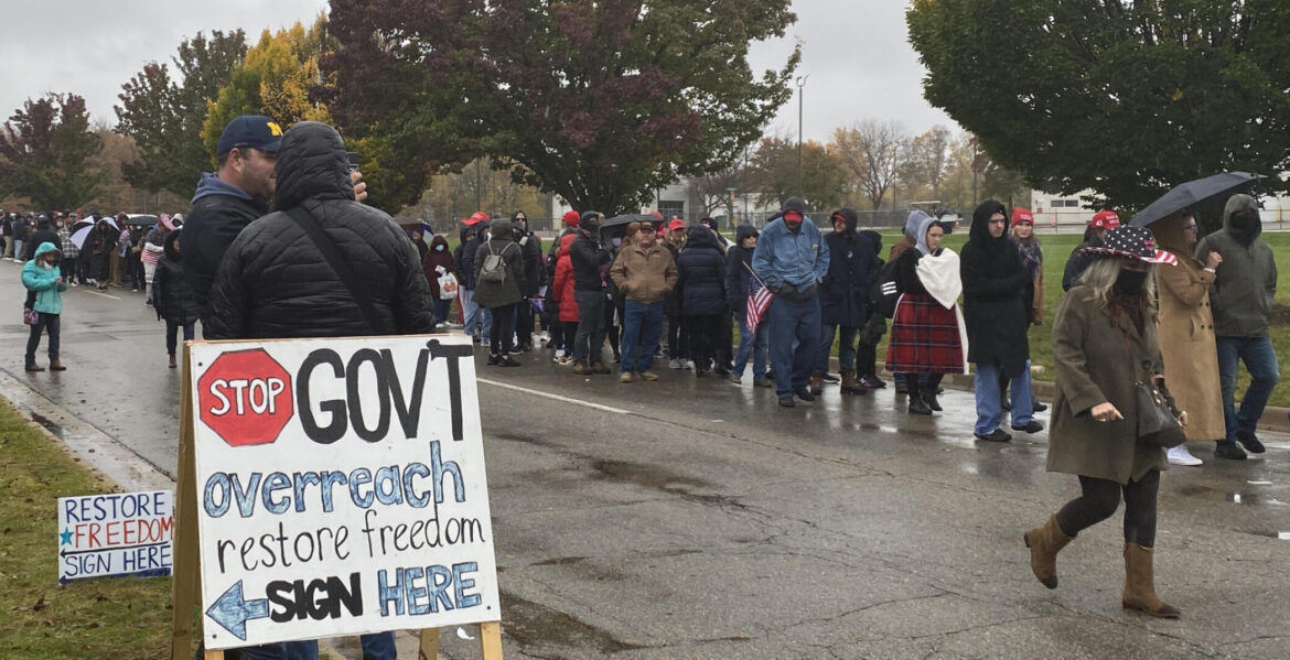 Line of people walk down sidewalk past a sign about government outreach.