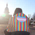 A person covering in a rainbow blanket sites in front of the Michigan Capitol
