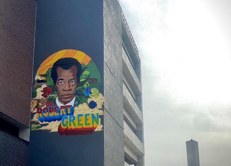 A colorful mural depicting a man’s portrait with the label “Dr. Robert Green” is painted on the side wall of a parking garage.