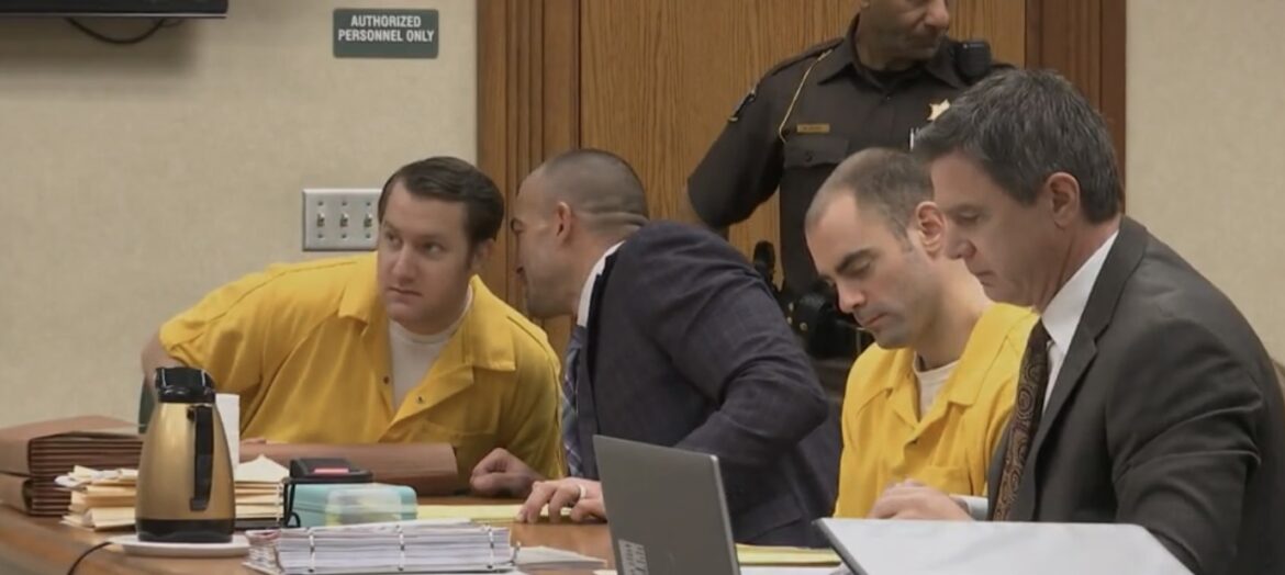 Two men in yellow jail clothes and their attorneys confer at courtroom table.